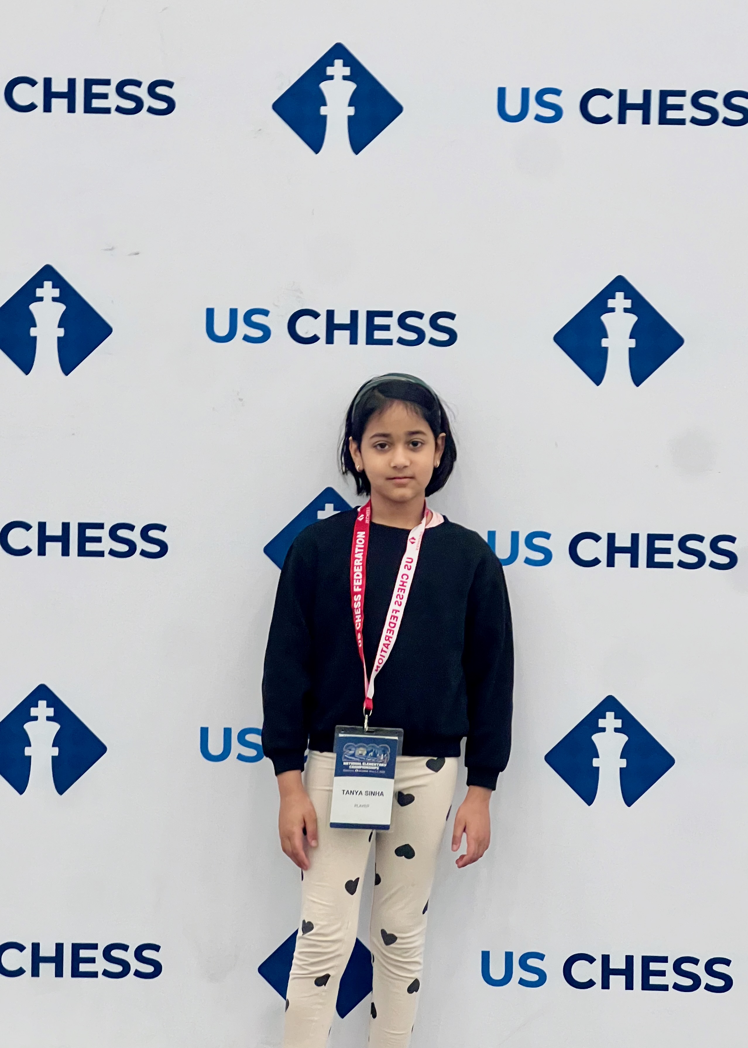 Tanya standing in front of the US Chess backdrop at the 2026 National Elementary Championships, wearing her player lanyard